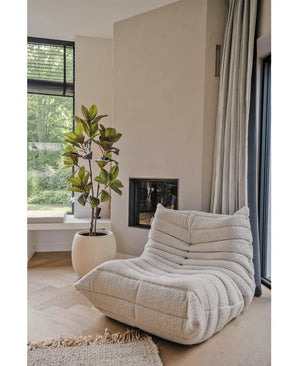 Een gezellige, crèmekleurige loungestoel met textuur staat bij een moderne open haard en een hoog raam. Ernaast staat de Kunstplant Yulaniaboom 150 cm in een ronde witte pot. Lichte houten vloeren en zacht zonlicht creëren een rustige, uitnodigende sfeer.