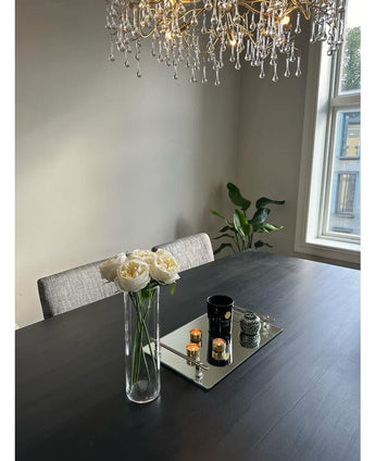 Een moderne eettafel met grijze stoelen, een Kunstrozenboeket "Carmen" Real Touch Crème met 36 cm boeket, kaarsen en decor op een spiegeltray. Erboven hangt een kroonluchter, met een raam en een potplant op de achtergrond.