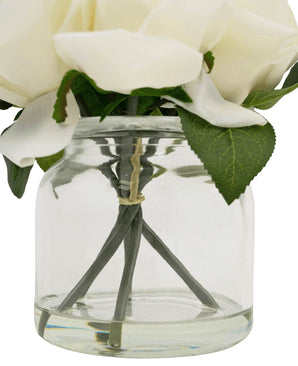 Een close-up van de Kunstrozenboeket Real Touch Wit 18cm: witte kunstbloemen met groene bladeren in een heldere glazen pot, zichtbare stelen bijeengehouden door een elastiekje.