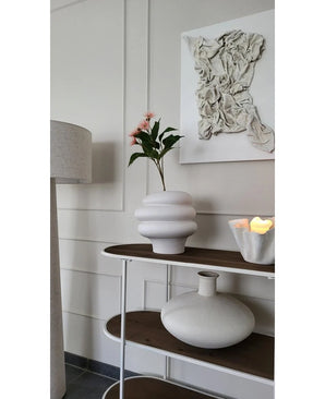 A modern interior with a white textured wall art, a white sculptural vase with Artificial Branch Flowering Eucalyptus 62 cm old pink, a decorative candle and a round ceramic vase on a wooden shelf next to a floor lamp.