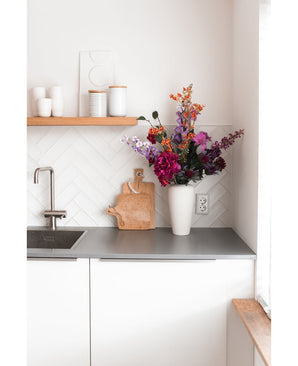 A modern white kitchen with a gray countertop, a sink, a wooden cutting board and a white vase filled with Artificial Flower Hydrangea 55 cm dark pink. White cups and pots stand on a wooden shelf above the counter.