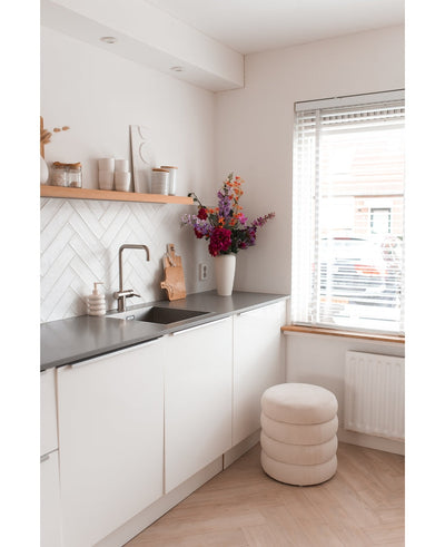 A modern, minimal kitchen with white cupboards, a black countertop and an Artificial Hydrangea flower 55 cm dark pink in a vase near the countertop, plus wooden shelves with decor and a round stool with cushions near a window with blinds.