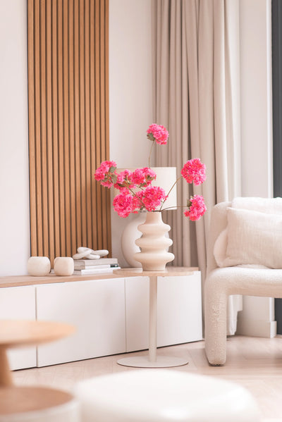 Een moderne woonkamer is voorzien van een witte bijzettafel met de Kunsttak Kersenbloesem 85 cm roze, waaraan felroze bloemen zijn toegevoegd. Een neutrale inrichting, lichte houtaccenten, beige gordijnen en een fauteuil met structuur maken de gezellige, minimalistische ruimte compleet.
