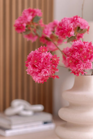 Een witte vaas van keramiek houdt een Kunsttak Kersenbloesem 85 cm roze vast. De achtergrond toont verticale houten panelen, een stapel boeken en een klein decoratief object bovenop.