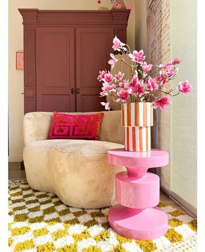 A cozy living room with a beige round sofa, a pink and orange geometric cushion, a pink side table, artificial branch Magnolia small flowered 89 cm pink in a vase, a yellow-white patterned rug and a chestnut brown cupboard in the background.