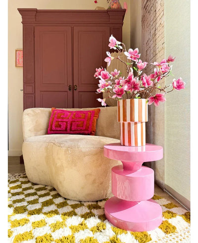 Een gezellige woonkamer met een beige ronde bank, een roze en oranje geometrisch kussen, een roze bijzettafel, Kunsttak Magnolia kleinbloemig 89 cm roze in een vaas, een geel-wit gedessineerd vloerkleed en een kast in kastanjebruin op de achtergrond.