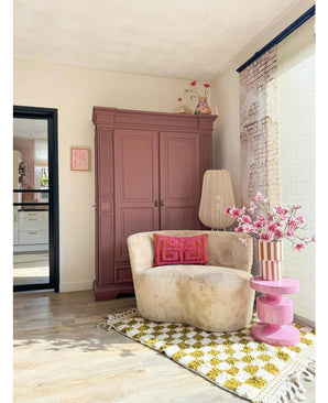Een gezellige leeshoek heeft een crèmekleurige stoel, roze kussens, een geel gedessineerd vloerkleed en een mauve kast. Op een roze bijzettafel staat de Kunsttak Magnolia kleinbloemig 89 cm roze, terwijl grote ramen deze uitnodigende ruimte vullen met zacht licht.