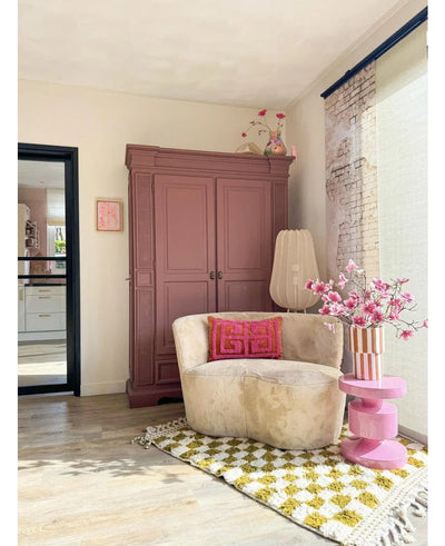 Een gezellige leeshoek heeft een crèmekleurige stoel, roze kussens, een geel gedessineerd vloerkleed en een mauve kast. Op een roze bijzettafel staat de Kunsttak Magnolia kleinbloemig 89 cm roze, terwijl grote ramen deze uitnodigende ruimte vullen met zacht licht.