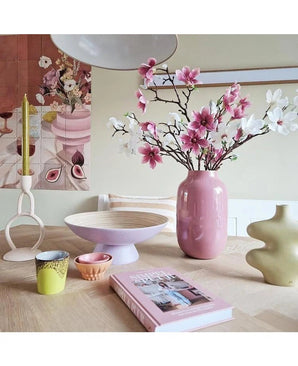 Op een lichte houten tafel staat de Kunsttak Magnolia kleinbloemig 89 cm wit in een roze vaas, modern keramiek, kleurrijke kopjes, een schaal, een gele kaars in een unieke houder en een roze boek, met bloemenkunstwerk op de muur erachter.