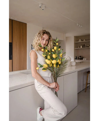 A woman with curly blonde hair, dressed in a cream-colored outfit, leans against a counter and holds the bouquet 'Artificial Branch Lemon 104 cm yellow', decorated with lush greenery. The modern kitchen is decorated in neutral colors and has wooden accents.