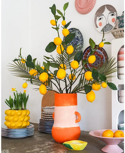 A bright room with a tall vase full of lemons and green leaves, in which the striking Kunsttak Lemon 104 cm yellow occupies a prominent place in the arrangement, surrounded by colorful ceramic bowls, a bowl of lemons and decorative plates on the wall - creating a cheerful, eclectic look.
