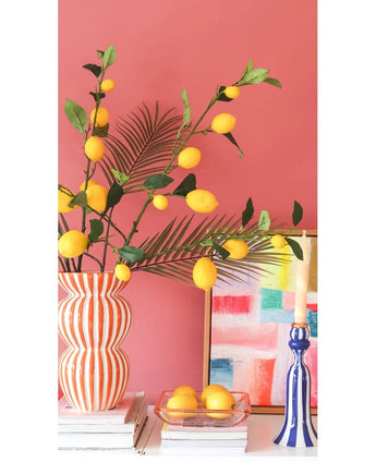 Against a coral pink wall are a striped vase filled with Kunsttak Lemon branches of 104 cm in yellow, a colorful painting, a stack of books, a blue candlestick with a candle and a tray of lemons.