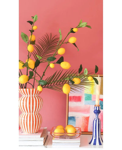 Against a coral pink wall are a striped vase filled with Kunsttak Lemon branches of 104 cm in yellow, a colorful painting, a stack of books, a blue candlestick with a candle and a tray of lemons.