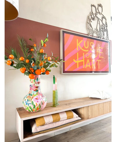 On a wooden shelf stands a colorful vase containing the Art Branch Mandarin 93 cm orange, next to a framed work of art with pink stripes that reads “Kissje op je Hart”. Also on the shelf are a green candle and folded blankets.