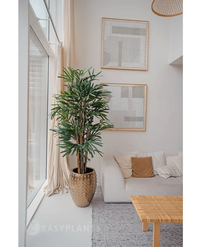 A Lady Artificial Palm 180 cm is placed near a large window in a bright, modern living room with a white sofa, beige cushions, woven table and abstract art. It adds a cozy minimalist style with its neutral tones and green artificial plant charm.