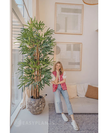 A woman in a pink patterned cardigan and jeans smiles on a beige sofa next to the Lady Artificial Palm 210 cm in a bright, modern living room with abstract art on the wall.