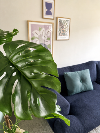 Een Kunstplant Monstera 120 cm staat prominent vooraan. Daarachter staat een donkerblauwe bank met een lichtblauw kussen tegen een beige muur met drie ingelijste kunstwerken.