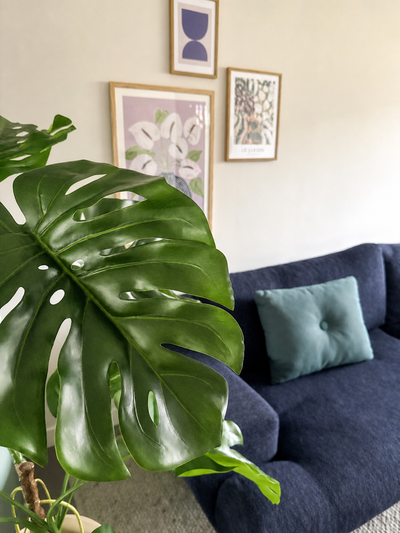 Een Kunstplant Monstera 120 cm staat prominent vooraan. Daarachter staat een donkerblauwe bank met een lichtblauw kussen tegen een beige muur met drie ingelijste kunstwerken.