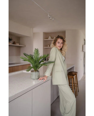 Een vrouw met krullend blond haar in een licht pak leunt tegen een keukeneiland, naast een Kunstpalm Baby Cycus 45 cm, die de neutrale inrichting van de moderne en minimalistische keuken opfleurt met stijlvolle kunstplant interieur flair.
