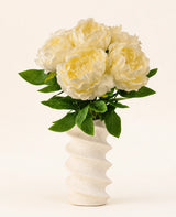 Een golvende, gestructureerde witte vaas bevat het Zijden kunstbloemen boeket Peony Wit-grote crèmekleurige pioen kunstbloemen met groene bladeren tegen een effen lichte achtergrond.