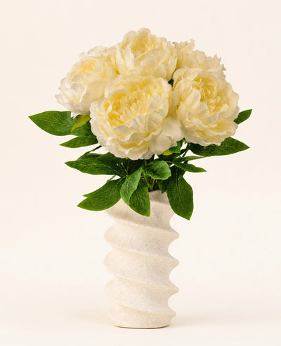 Een golvende, gestructureerde witte vaas bevat het Zijden kunstbloemen boeket Peony Wit-grote crèmekleurige pioen kunstbloemen met groene bladeren tegen een effen lichte achtergrond.