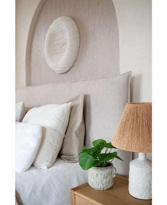 Een knusse slaaphoek met een beige hoofdeinde, witte en beige kussens, een houten nachtkastje, een keramische lamp met een geweven kap, de Kunst Pannenkoekplant 27 cm en witte muurkunst boven het bed.