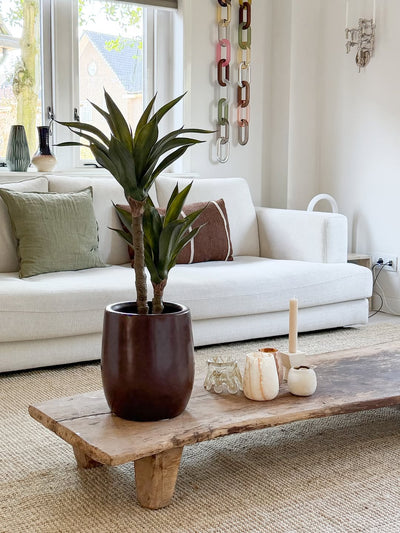 Een moderne woonkamer met een witte bank, groene en bruine kussens, een houten salontafel versierd met kaarsen en de Kunstplant Agave 76 cm, en een wanddecoratie van kettinggaas. Door de ramen achter de bank valt natuurlijk licht naar binnen.