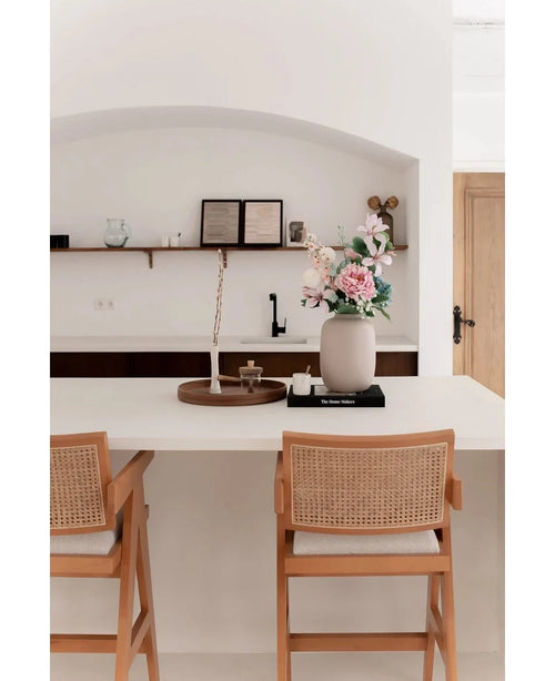 A minimalist kitchen has a white island with two wooden chairs and the Vase Artic Beige Ø21 x H29 cm with pink and white flowers. Shelves with books and ceramics are attached to the wall in the background.