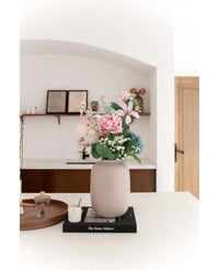 De Vaas Artic Beige Ø21 x H29 cm, gevuld met roze en blauwe kunstbloemen, staat op een zwart boek op een wit aanrecht. Zachte verlichting accentueert het minimalistische tafereel met decorplanken en een houten deur op de achtergrond.