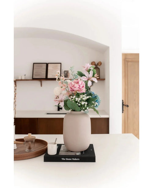 The Vase Artic Beige Ø21 x H29 cm, filled with pink and blue artificial flowers, stands on a black book on a white countertop. Soft lighting accentuates the minimalist scene with decor shelves and a wooden door in the background.