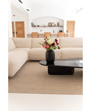 Een moderne woonkamer met een beige sofa, neutraal vloerkleed en een zwarte salontafel met de Vaas Artic Black Ø21 x H29 cm met roze, witte en rode bloemen. Op de achtergrond verschijnen planken en houten deuren.