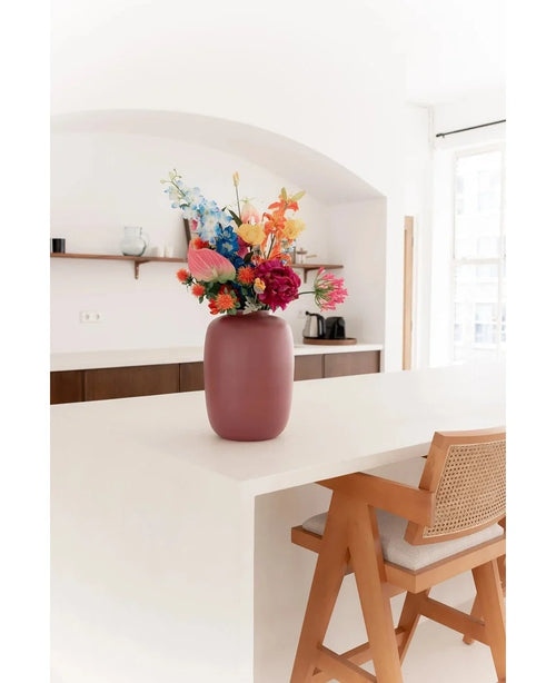 The Artic Pink Vase Ø21 x H29 cm, filled with colorful flowers, stands on a white kitchen island next to a wooden chair with a woven back. Minimalist shelves and a coffee maker adorn the bright, naturally lit space.