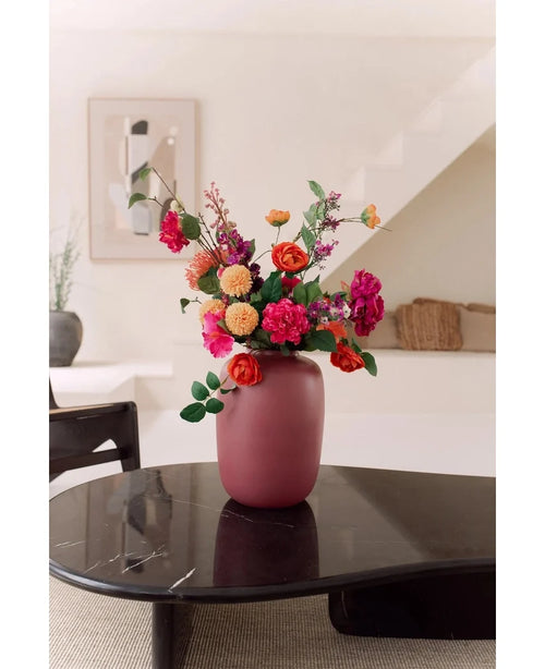 The Vase Artic Pink Ø21 x H29 cm, filled with pink, orange and red flowers, stands on a dark glossy coffee table in a modern minimalist living room with a staircase and framed works of art in the background.
