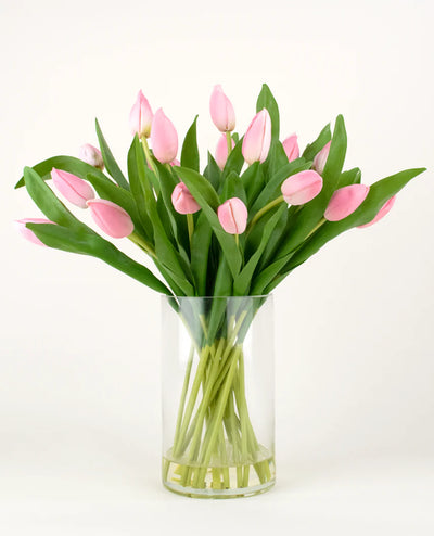 Het Zijden kunstboeket Tulpen in vaas Roze heeft roze tulpen met lange stelen en groene bladeren in een heldere glazen vaas, tegen een effen witte achtergrond.