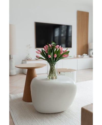 Een moderne woonkamer met een wit vloerkleed, een grote ovale salontafel en een kleine ronde houten bijzettafel. Op de tafel staat het Zijden kunstboeket Tulpen in vaas Roze; een tv en een minimalistisch decor maken de achtergrond compleet.