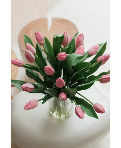 Het Zijden kunstboeket Tulpen in vaas Roze toont roze kunsttulpen met groene bladeren, gerangschikt in een glazen vaas, tentoongesteld op een wit vlak met een zacht vervaagde lichte achtergrond, van bovenaf gezien.