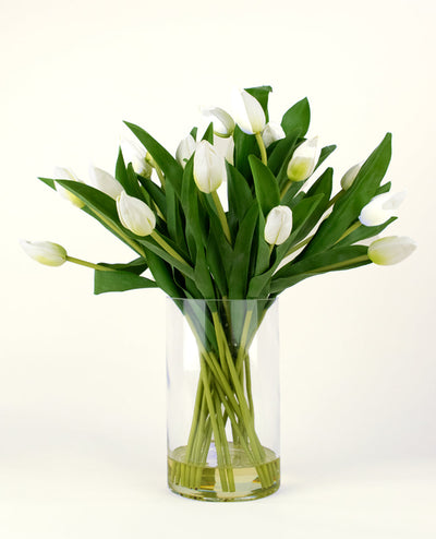 Het Zijden kunstboeket Tulpen in vaas Wit heeft witte tulpen met groene bladeren en stelen in een heldere glazen vaas, tegen een eenvoudige lichte achtergrond.